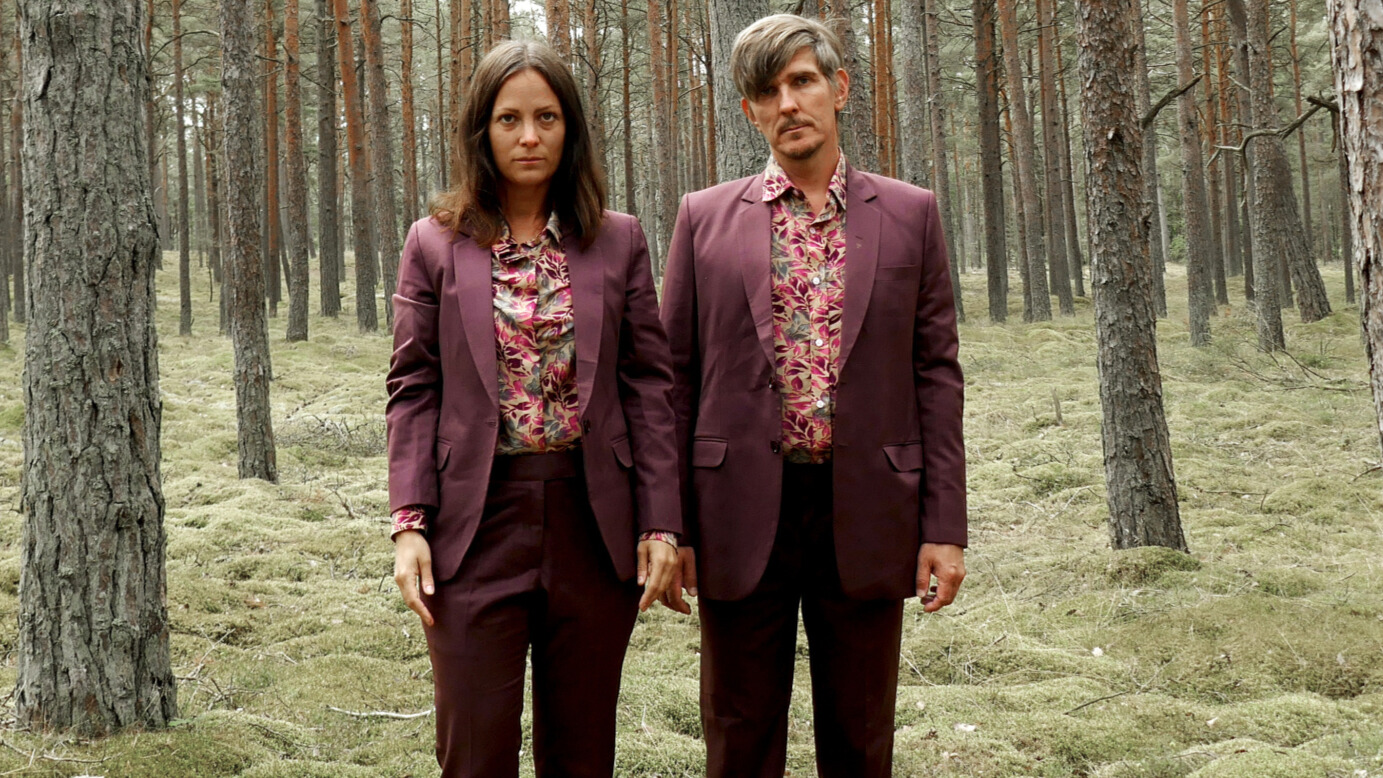 Två konstnärer i vinröda kostymer står i en skog